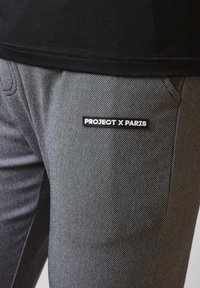 Grå teksturerede bukser med et diskret mønster. Har sorte sidelæg og et "PROJECT X PARIS" mærke i et rektangulært design.