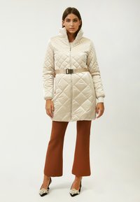 Abrigo acolchado largo de color crema con un alto cuello, cintura ceñida y puños acanalados, combinado con pantalones marrones de campana y tacones negros.