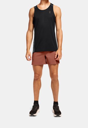 Mężczyzna ubrany w czarną sportową koszulkę bez rękawów, rdzawe spodenki, czarne skarpetki i czarne buty do biegania, stojący na jednolitym tle.