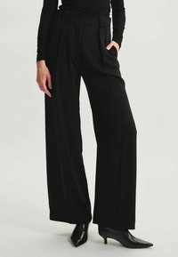 Pantalons larges noirs à taille haute, avec plis, deux poches avant et fermeture à deux boutons. Texture lisse avec une coupe ajustée.