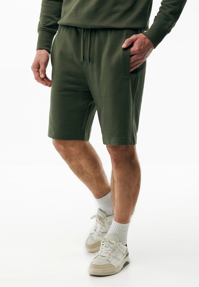 Man draagt olijfgroene shorts met trekkoord, witte sportsokken en witte sneakers, staand met één hand in de zak.