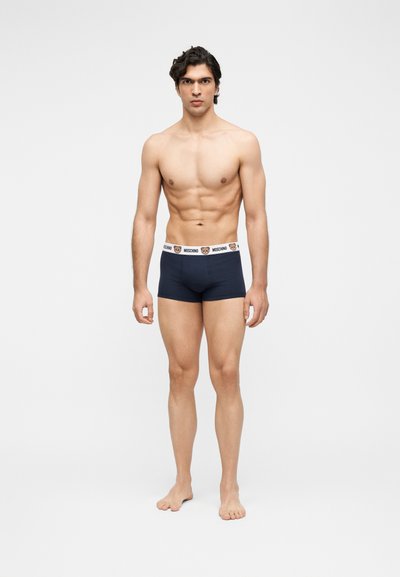 Μυώδης άνδρας που στέκεται ξυπόλητος σε λευκό φόντο, φορώντας ναυτικό μπλε μποξεράκια με λευκή μέση που έχει αρκουδάκι και το κείμενο "Moschino".