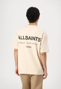 Beige oversized t-shirt made of cotton, featuring a large black "ALLSAINTS" logo and text on the back. Short sleeves and crew neckline.