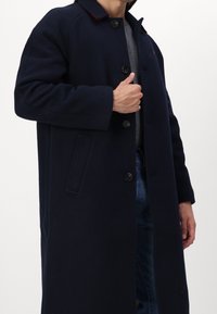 Cappotto di lana blu navale con chiusura a bottoni, grandi tasche e un colletto rosso a contrasto. Il tessuto ha una finitura morbida e testurizzata.
