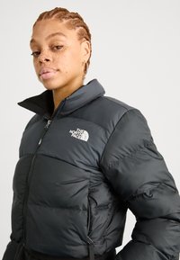 Černá péřová bunda s vysokým límcem, se zipem na přední straně a logem North Face na levé hrudi; prošívaný design, zateplený materiál.