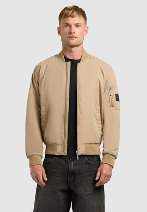 Férfi világosbarna bomberdzsekit visel fekete póló és sötét farmer fölött, egyszínű világosszürke háttér előtt áll.