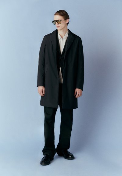 Lang sort frakke over en mørk vest, parret med en lys skjorte og mørke bukser. Slanke sorte sko og oversized grønne solbriller.