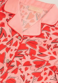 Haut de pyjama en satin rose avec motifs de cœurs rouges, texte manuscrit noir, passepoil rouge sur le col et la poche, et deux boutons argentés.