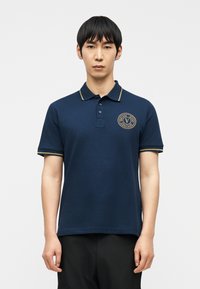 Νεαρός άνδρας που στέκεται, φοράει μπλε σκούρο πόλο με χρυσή λεπτομέρεια και κυκλικό λογότυπο «Versace Jeans Couture» στο στήθος, μαύρο παντελόνι.