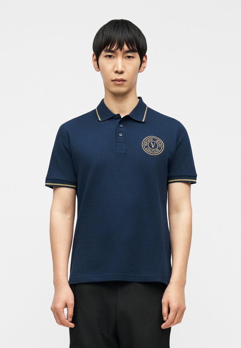 Νεαρός άνδρας που στέκεται, φοράει μπλε σκο ύρο πόλο με χρυσή λεπτομέρεια και κυκλικό λογότυπο «Versace Jeans Couture» στο στήθος, μαύρο παντελόνι.