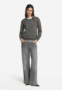 Kobieta w szarej długiej bluzie z długim rękawem i szerokich, wyblakłych szarych dżinsach, z rękami w kieszeniach, stojąca na białym tle.