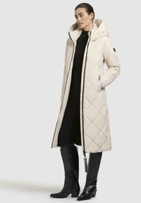 Cappotto lungo imbottito beige con cappuccio, dotato di una cerniera frontale, tasche laterali e un logo a contrasto nero. Indossato con un abbigliamento nero aderente e stivali alti.