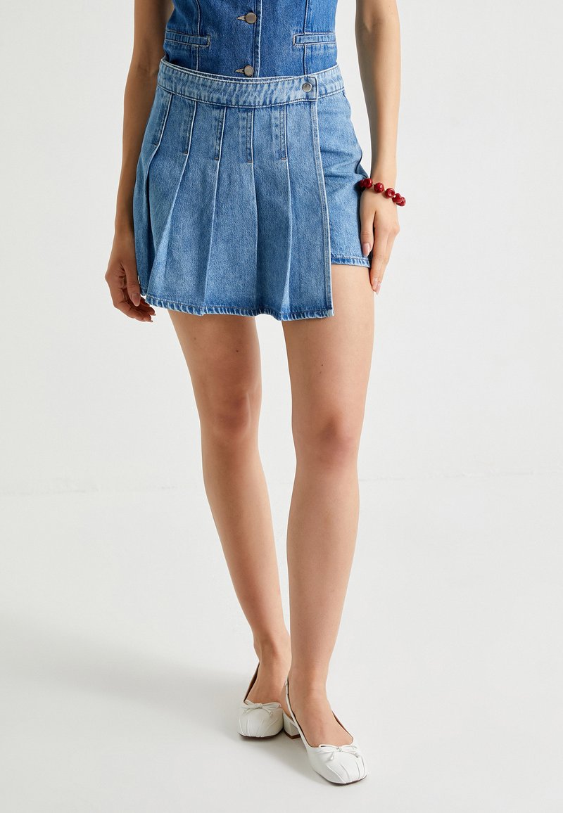 Falda de mezclilla con pliegues, color azul, diseño de cintura alta, dobladillo sin terminar. Combinada con zapatos blancos. Textura suave, estilo casual.