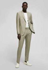 Costume beige clair avec une coupe slim, présentant des revers en angle et deux boutons. Associé à une chemise blanche à col rond et des mocassins blanc cassé.