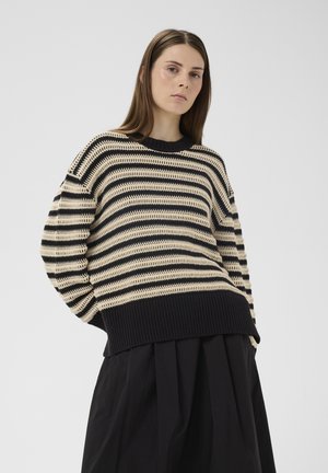 Žena v béžovom a čierno pruhovanom pletenom svetri a čiernej plisovanej sukni stojaca pred jednofarebným svetlým pozadím.