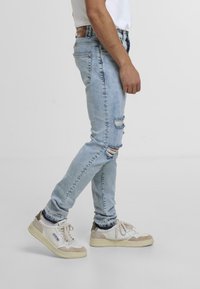 Heledad hall kuni sinised, kulunud teksapüksid, millel on rebenenud laigud ja kitsas lõige, koos beežide ja valgete ninatossudega.