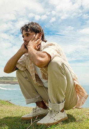 Hombre con una camisa de cuadros de manga corta, pantalones crema y zapatillas blancas, agachado en la hierba con las manos en la cara. Cielo y océano al fondo.