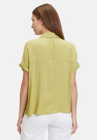 Short-sleeve, light green blouse with a curved back yoke, relaxed fit, and rolling cuffs, made from soft, lightweight fabric.