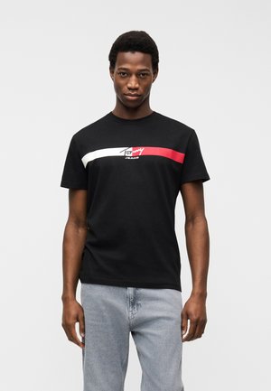 Jeune homme portant un t-shirt noir Tommy Jeans avec des rayures horizontales blanches et rouges, associé à un jean bleu clair, debout devant un fond uni.