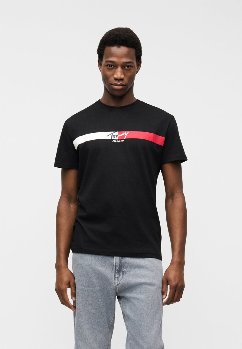 Jeune homme portant un t-shirt noir Tommy Jeans avec des rayures horizontales blanches et rouges, associé à un jean bleu clair, debout devant un fond uni.