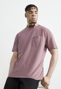T-shirt à manches courtes, couleur rose, en coton, avec une poche poitrine dotée d'un logo carré. Design minimaliste et texture douce.