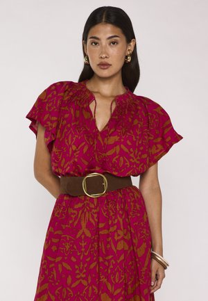 Donna con capelli lunghi e scuri che indossa orecchini a cerchio dorati e un vestito rosso a motivo con maniche arricciate e una cintura larga marrone con fibbia dorata.