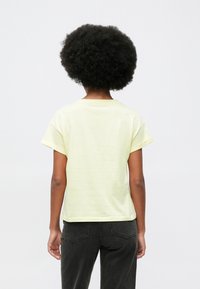 Άτομο με αφρο μαλλιά φοράει ανοιχτό κίτρινο κοντομάνικο μπλουζάκι και μαύρο τζιν, στέκεται με πλάτη προς το φακό σε απλό ανοιχτό φόντο.