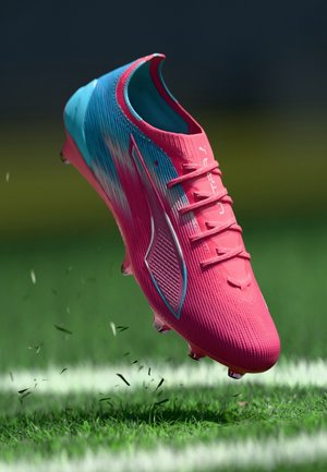 Pink-blauer Fußballschuh mit Markenlogo, Schnürsenkeln und Stollen, der Gras auf einem markierten Sportplatz hochwirbelt.