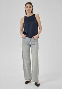 Marine satijnen mouwloze top gecombineerd met lichtblauwe high-waisted jeans; model staat met handen in de zakken en draagt zwarte hakken.