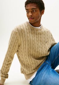 Beige kabelgebreide trui met een ronde hals en geribbelde manchetten. Heeft een klein logo op de linkerborst. Gemaakt in combinatie met blauwe jeans.