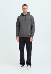 Felpa grigia con tasca a marsupio, cappuccio con cordoncino e piccolo logo sul petto; abbinata a pantaloni neri e sneaker bianche.