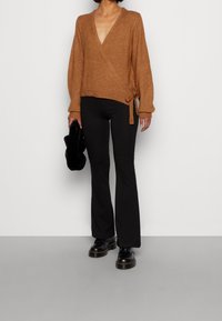 Brun innesvärmd tröja med djup V-ringning, texturerad stickning och knytning vid midjan, parat med åtsittande svarta utsvängda byxor och svarta skor.