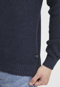Tengerész kék kötött pulóver texturált mintával, bordás mandzsettákkal és címkével az alján. Az anyaga lágy és közepes súlyú.