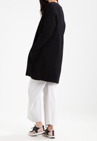 Černý dlouhý vlněný kardigan s uvolněným střihem, spárovaný s bílými kalhotami s širokými nohavicemi a multibarevnými teniskami s možností šněrování.