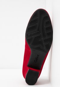Chaussure plate en daim rouge montrant une semelle en caoutchouc noire texturée avec la marque Gabor et la taille 4½ visible sur un fond blanc.