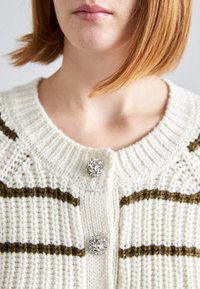 Stickad cardigan med vita och bruna ränder, med rund halsringning och två utsmyckade knappar med blommönster fram.