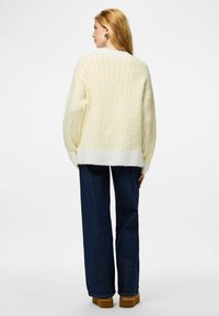 Šviesiai geltonas megztas megztinis su tekstūruotu raštu, su ribotu apvadu ir platėjančiomis rankovėmis, derinamas su tamsiai mėlynomis plačiomis kojinių džinsais.