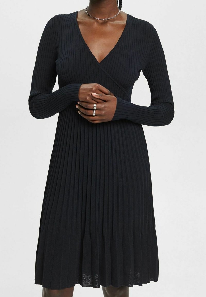 Femme portant une robe noire en maille côtelée à manches longues, avec un col en V et une jupe plissée, accessoirisée avec des bagues et un collier chaîne.