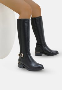 Botas de cuero negro hasta la rodilla con un detalle de correa y hebilla, cremallera lateral y un tacón bajo tipo bloque. Textura suave con acentos mínimos.