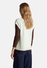 Weißer gestrickter Weste mit geripptem Saum, getragen über einem braunen Langarmshirt. Die Rückansicht zeigt die glatte Textur und die lockere Passform. Dunkelblaue weit geschnittene Jeans.