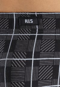 Schwarzer Stoff mit einem strukturierten, geometrischen Muster in verschiedenen Farbtönen. Mit einem Etikett, das "H.I.S." am Bund zeigt.