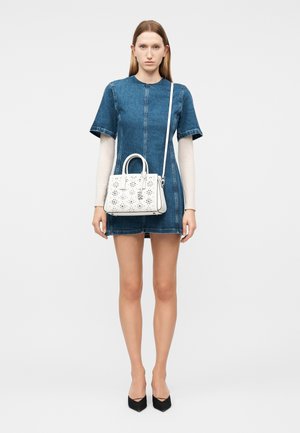 Ženska v kratki jeans obleki in belih dolgih rokavih drži belo torbico s cvetličnim vzorcem in naramnim trakom, stoji pred enobarvnim ozadjem.