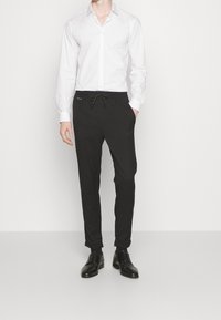 Pantalons noirs à cordon de serrage avec une coupe slim, en matériau lisse. Associés à une chemise blanche à manches longues et des chaussures de ville noires.