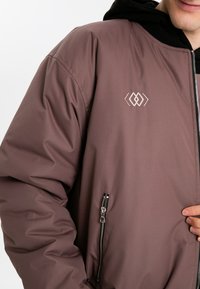 Lätt mauve jacka med en dragkedjeförsedd sidoficka och broderad logotyp. Har en slät textur och en avslappnad passform.