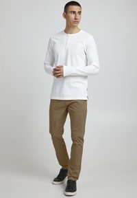 Langermet hvit henley-skjorte med knapper, kombinert med lysebrune bukser og svarte joggesko. Avslappet passform, jevn tekstur i stoffet.