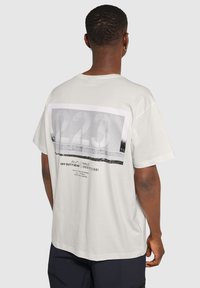 Ljusgrå kortärmad t-shirt med ett grafiskt tryck på baksidan som har stora siffror och text i svart och vitt. Avslappnad passform.