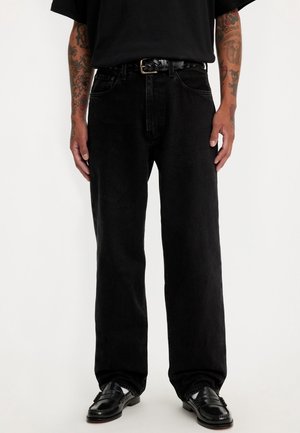 Homme portant un jean noir ample avec une ceinture noire tressée, des chaussures noires et des bras tatoués sur un fond uni.