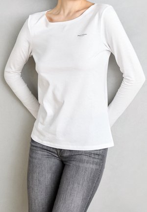 Persoon draagt een effen wit shirt met lange mouwen met het logo "Marc O'Polo" op de borst, gecombineerd met een grijze spijkerbroek tegen een neutrale achtergrond.