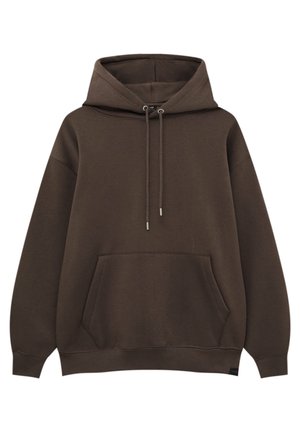 Jeune personne portant un sweat à capuche marron, un jean bleu large et des baskets blanches, debout, de dos sur un fond clair uni.