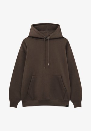 Ung person iført en brun hættetrøje, løse blå jeans og hvide sneakers, stående med ryggen mod en ensfarvet lys baggrund.